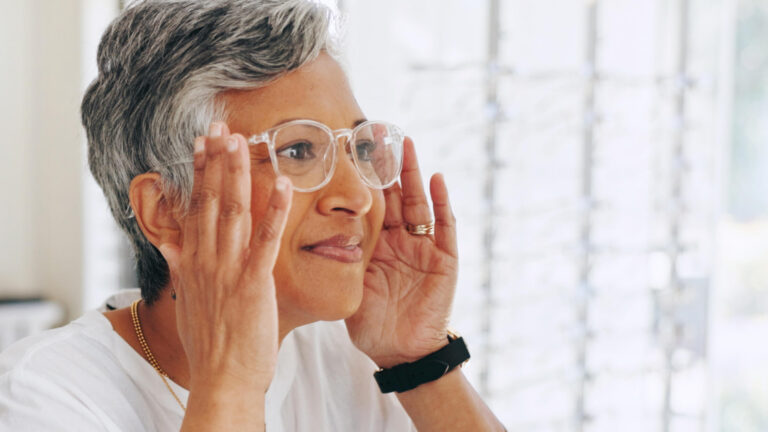 Middle-aged woman with short gray hair trying on clear-framed glasses at an optical shop, with eyeglass frames displayed on the wall behind her