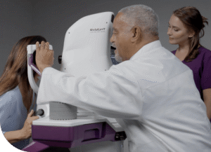 Doctor using an RxSIGHT Light Delivery Device to administer a UV light treatment to a female patient's eye, with a medical assistant observing in the background