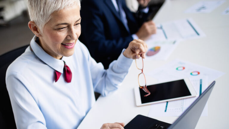 Smiling businesswoman holding her eyeglasses away from her face while working on a laptop in an office setting, representing clear vision after cataract surgery.